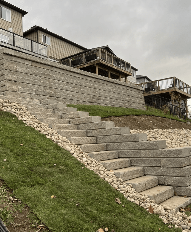 Terraced backyard with stone retaining walls and steps, bordered by grass and rocks. Residential houses and wooden decks are visible above.